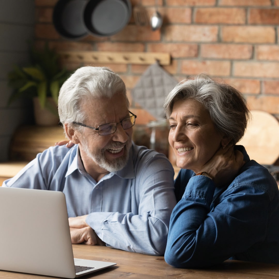 A couple with gray hair in their kitchen with a brick wall behind them, sitting at the counter together with a laptop experiencing the benefits of online couples counseling.