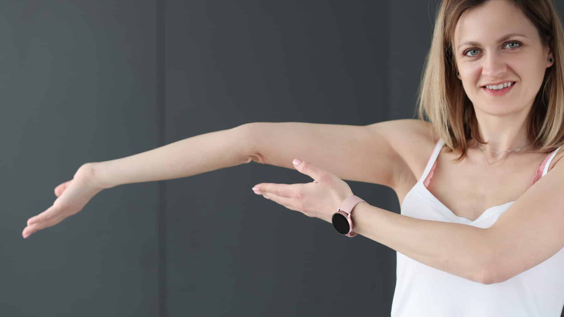Young woman with shoulder length blond hair wearing a white camisole standing in front of  a dark gray wall demonstrating her flexibility for Ehlers-Danlos Syndrome (EDS) and Hypermobility Awareness Month by hyperextending her right elbow.