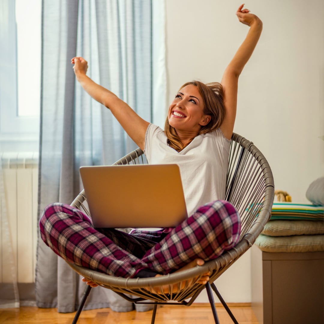 Young woman wearing red plaid pajama pants and sitting cross legged in a round string chair whith a laptop stretching her arms out revealing her hypermobile elbows for Ehlers-Danlos Syndrome and Hypermobility Awareness Month.