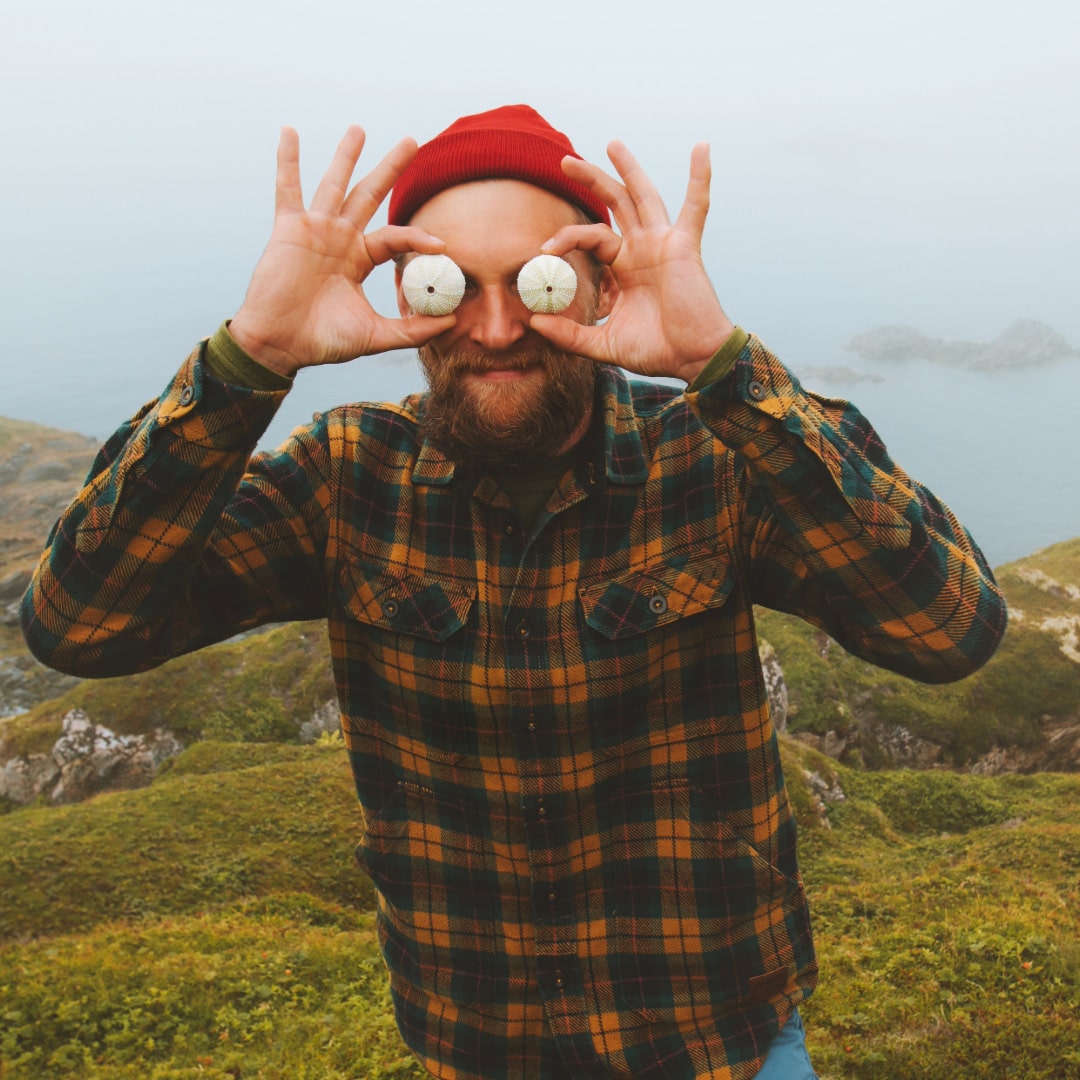 Man with a beard wearing a red beanie and a plaid flannel shirt near the shore embracing self-acceptance and authenticity by being silly holding up urchin skeletons over his eyes.