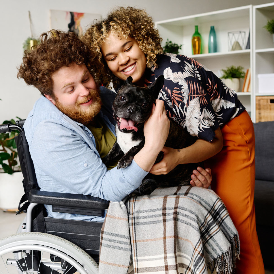 Man in a wheelchair and his partner enjoying their therapy dog at home together after family counseling for caregiving.