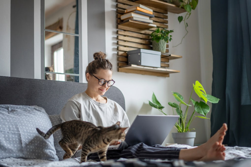 Middle aged woman with her hair up in a messy bun wearing glasses, a rumpled tee-shirt and loose striped pants sitting on a gray futon with her tabby cat and a laptop with her feet up working on finding the best therapist for EDS to get support for her chronic pain.