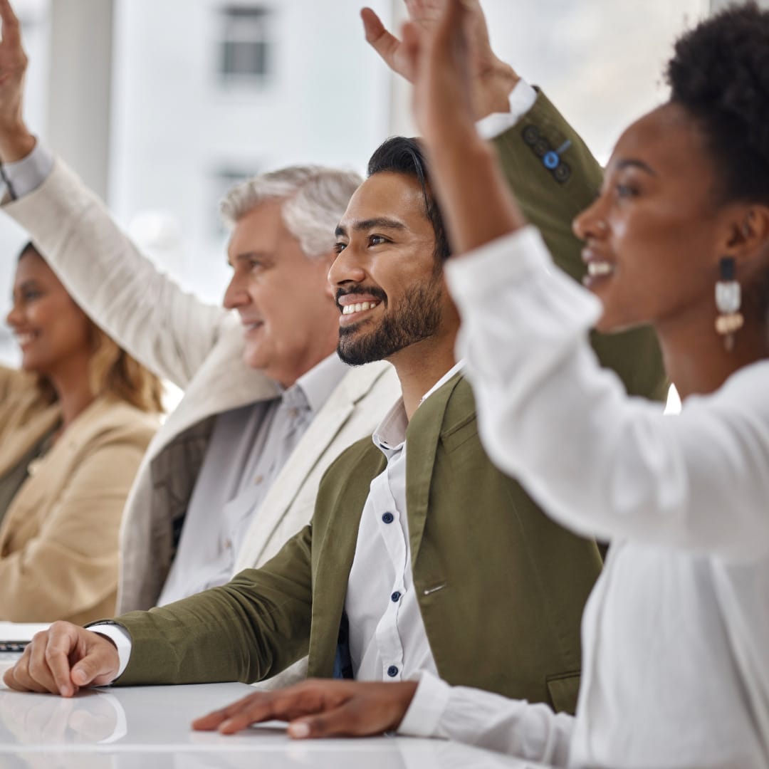 A row of adults in a class sitting at a table raising their hands to ask or answer the question, "What are the common symptoms of ADHD in adults?" The adults in the photo are, from front to back: A Black woman with her hair up wearing dangling earrings and a white blouse, a Black man with a beard wearing a green jacket over a white button up, an older white man with gray hair wearing a tan jacket over a white button up, and a Black woman with shoulder-length hair with blond highlights wearing a tan sweater.