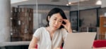 Asian woman wearing an off-white top, sitting in front of her laplaptop,lding her head with her left hand, looking frustrated and confused about finding ADHD therapy for adults.
