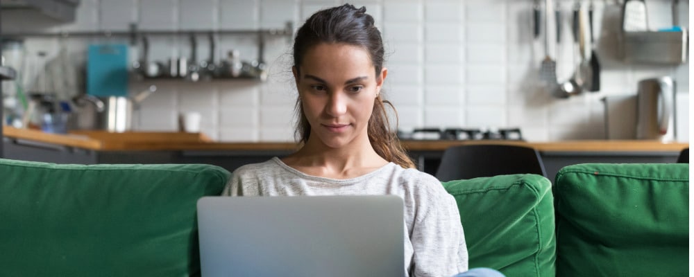 A woman with long dark hair pulled back wearing a white top sitting on a green couch with her kitchen in the background searching for an out-of-network therapist online.