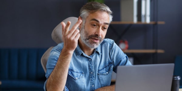 Man in  blue button down shirt with rolled up sleeves, with gray hair and beard gesturing to his laptop while receiving online anxiety treatment california.