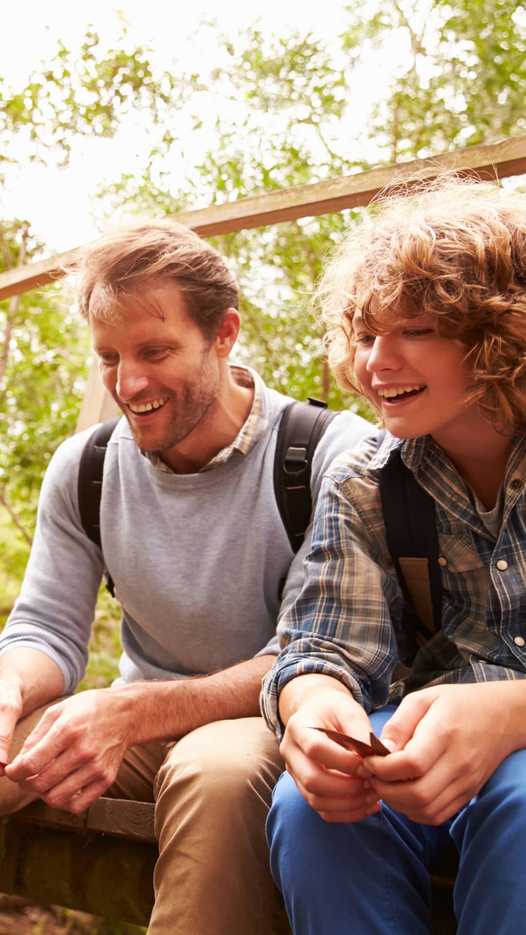 A father and son wearing backpacks sitting close together in nature smiling and experiencing an authentic connection.