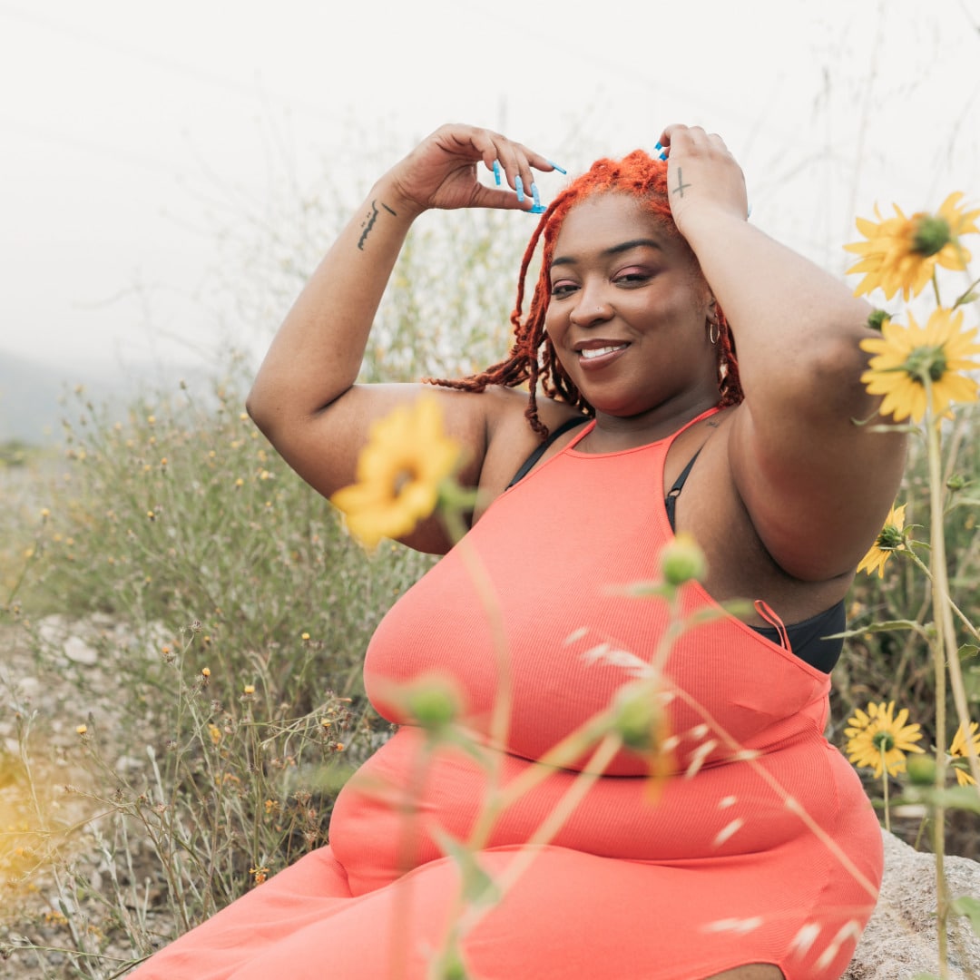 A Black woman in an orange dress sitting on a rock in a meadow reaping the benefits of self-love and recognizing inner worth while putting flowers in her hair.