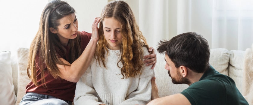 Mom sits on the couch with daughter while Dad crouches on the other side of her, as they provide support together using what the best online parenting counseling helped them understand.