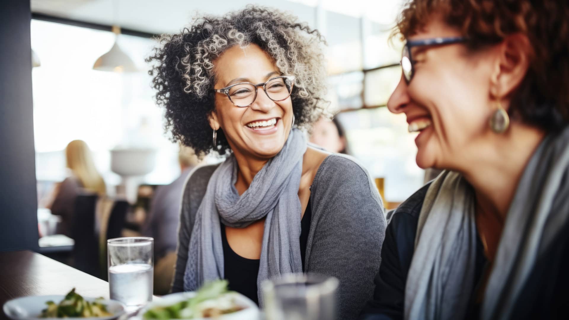 Two over-40 women laughing, one with curly gray hair and the other in the foreground with short dark hair, are sitting at a lunch counter together, building a strong support network over lunch. Both are wearing gray long-sleeve shirts, gray scarves, glasses, and earrings. They have been served healthy lunches with glasses of water.