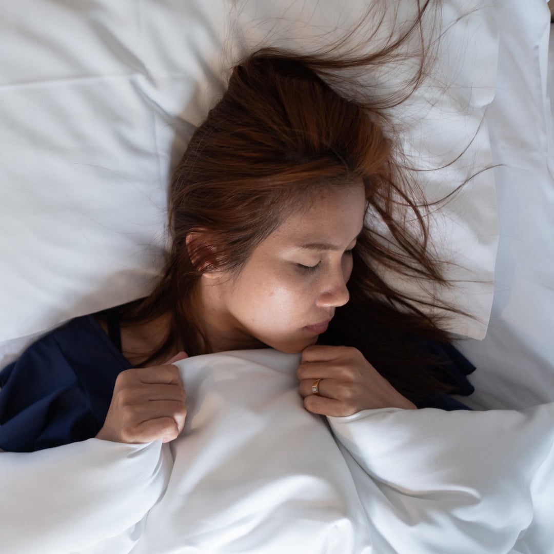 Asian woman with her hair whisping out on the pillow peacefully sleeping in a bed with white sheets while holding the top sheet with both hands practicing coping strategies for managing anxiety.