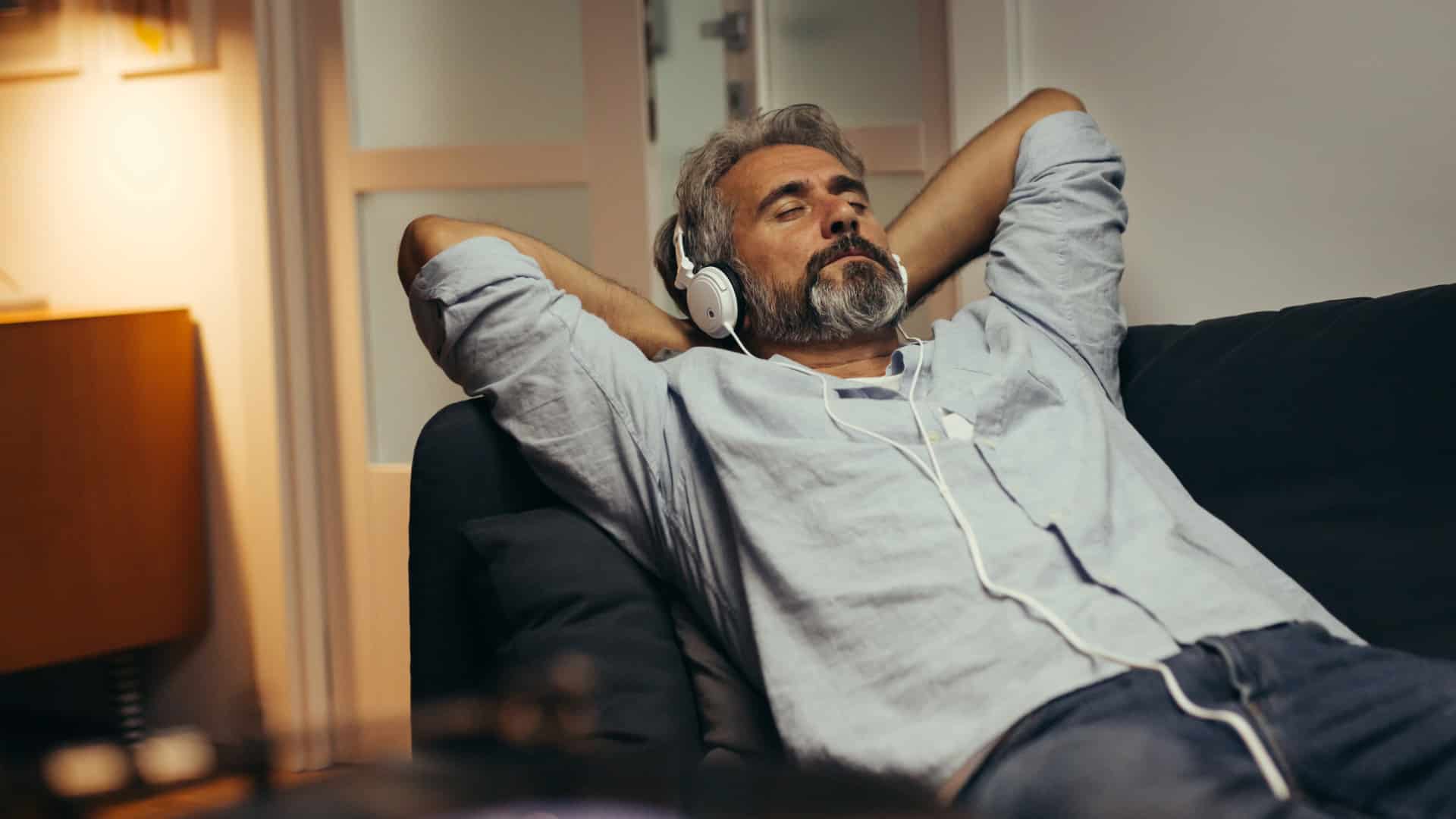 Man with salt and pepper hair and beard wearing a light gray button down shirt with the sleves rolled up laying back on a black couch with his hands behind his hands and elbows out listening to music through white headphones  demonstrating one of the coping techniques for adult PDA.