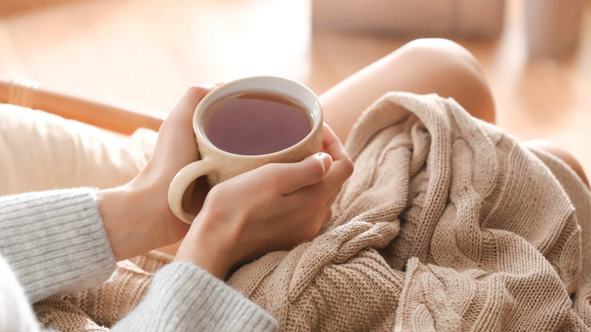 A woman sitting in a tan chair with a tan knitted blanket holding a cup of tea as self-care after counseling for chronic illness anxiety.