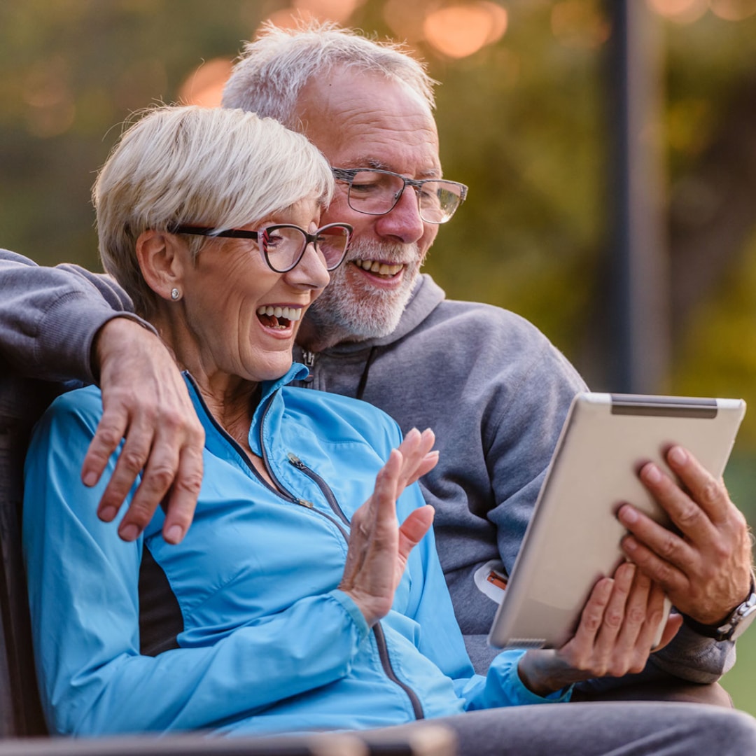 Couple, both with gray hair and glasses, sitting on a bench in their yard working with their counselor in california using a tablet.    