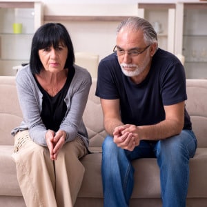 A middle-aged married couple sitting on the couch not touching in disagreement with their hands in their laps gazing downward to show difference between marital and premarital counseling.