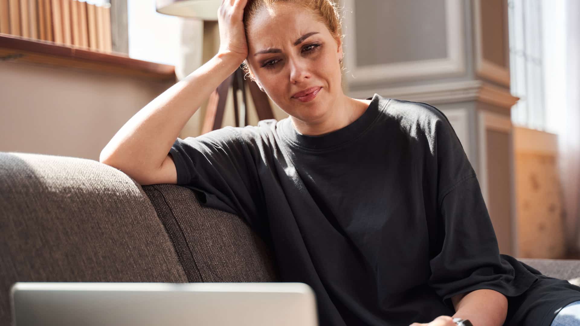 Middle aged woman with red hair pulled back wearing a charcoal gray crew neck t-shirt sitting on a tan couch with her laptop talking to  an experienced chronic illness therapist for dual diagnosis of anxiety and chronic illness.