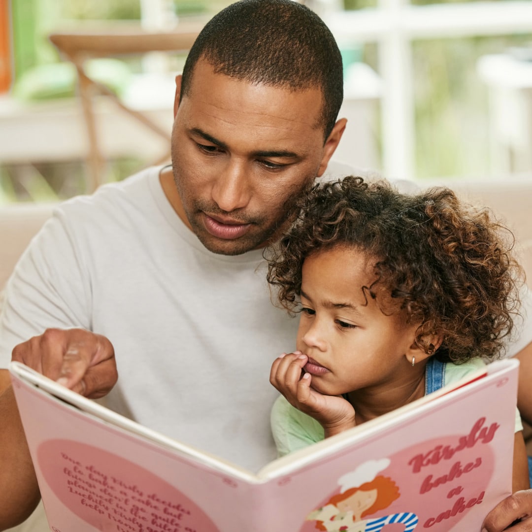 A Black father with his young daughter on his lap reading her a story while pointing to a page to support her healthy child development.