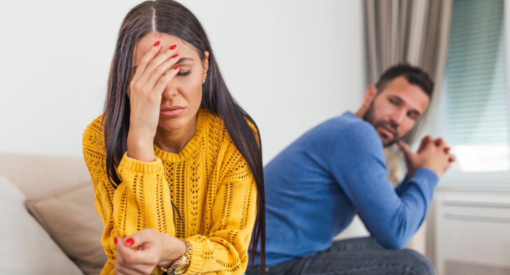 A couple sitting on opposite ends of the couch. The woman has her head in her hand with a sad, exasperated look on her face. The man is looking at her seriously with his hands clasped and his elbows on his knees, wondering how they can agree about individual child's needs.