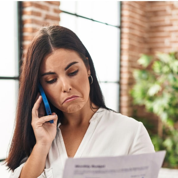 A woman with long dark hair wearing a white blouse standing in her living room on the phone  frowning while dealing with a denial to use insurance to pay for therapy.