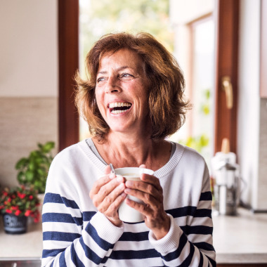 lifestyle adjustments for adhd control A woman in her 50s wearing a white sweater with navy blue stripes holding a cup of tea with both hands and enjoying lifestyle adjustments for adhd control she has made to calm her nervous system.
