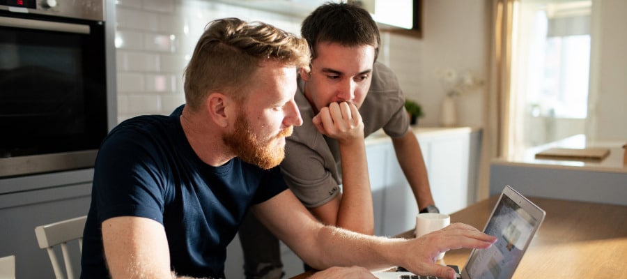 Gay couple in the kitchen working on mental health services reimbursement for therapy on their laptop. In foreground is a man with red hair and beard wearing a navy blue t-shirt using the laptop and behind him is a man with short dark hair and a khaki t-shirt leaning his elbow on the table and leaning his chin on his hand.