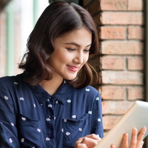 Young woman with short, dark hair wearing a navy blue button up shirt with a small white paisley design using a tablet to access neurodiversity psychological services.