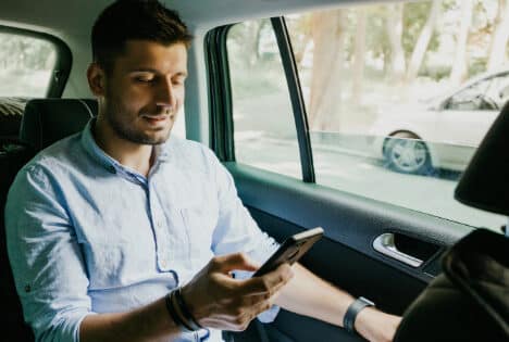 White man with dark hair and beard in backseat of his car, wearing a light blue long sleeve button up shirt, meeting with his therapist for online addiction counseling.