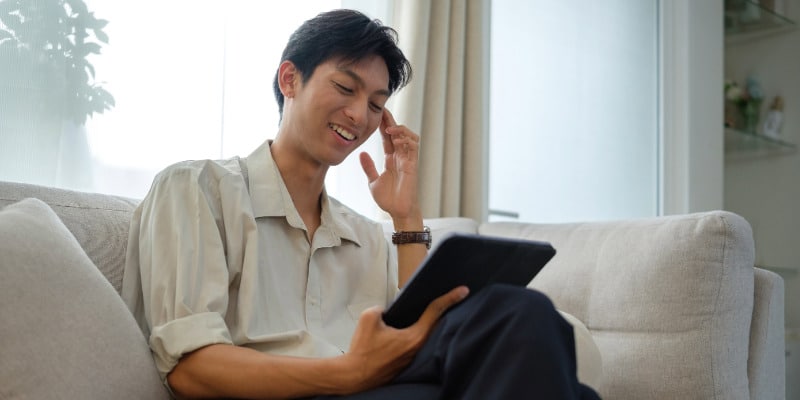 A young Asian man smiling and wearing a light tan button-up shirt with rolled up sleeves, sitting on an off-white couch, meeting with his therapist on his tablet for online ADHD treatment for adults.