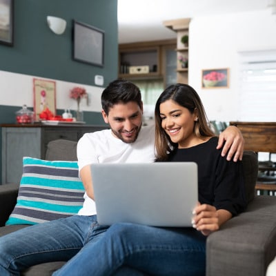 A smiling couple participating in online premarital counseling sitting closely on the couch sharing a laptop.