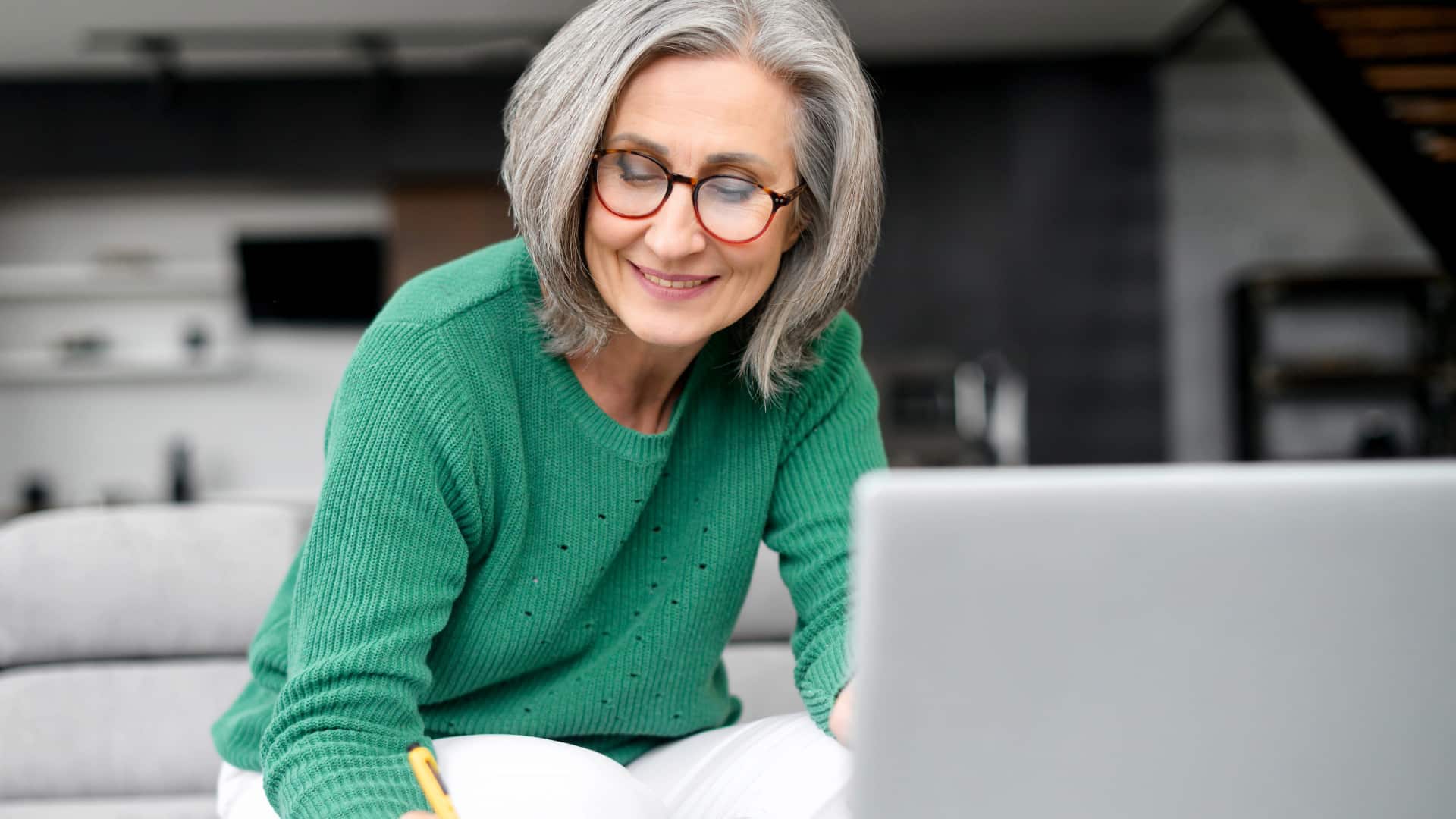 An experienced therapist with shoulder-length gray hair wearing a green sweater and white pants sitting in front of her laptop providing online private therapy for sobriety in her home office. 