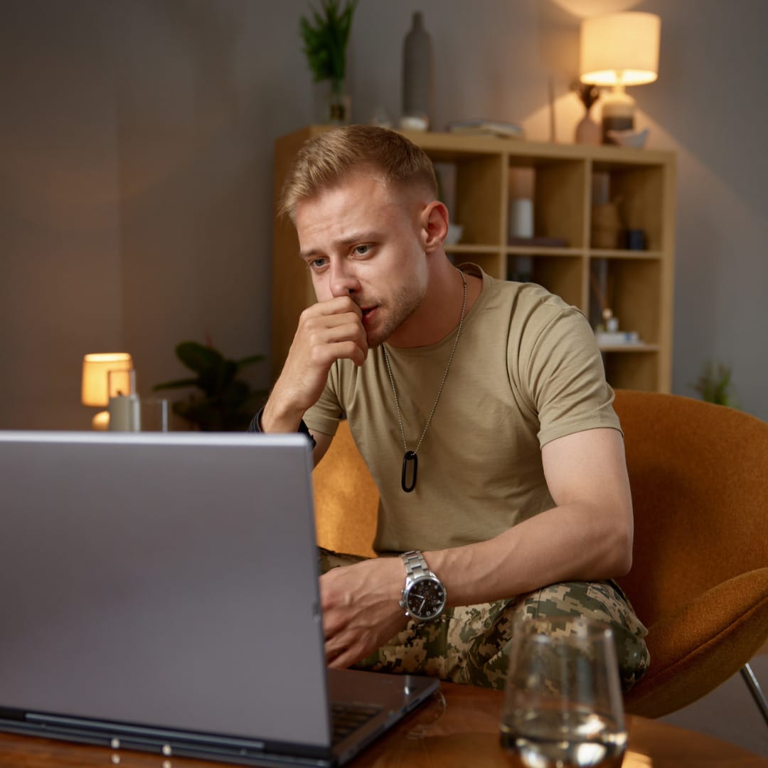 Man with short read hair wearing camo pants and a tan t-shirt sitting on a rust-colored chair in front of a laptop in his living room speaking to his therapist for online trauma treatment.