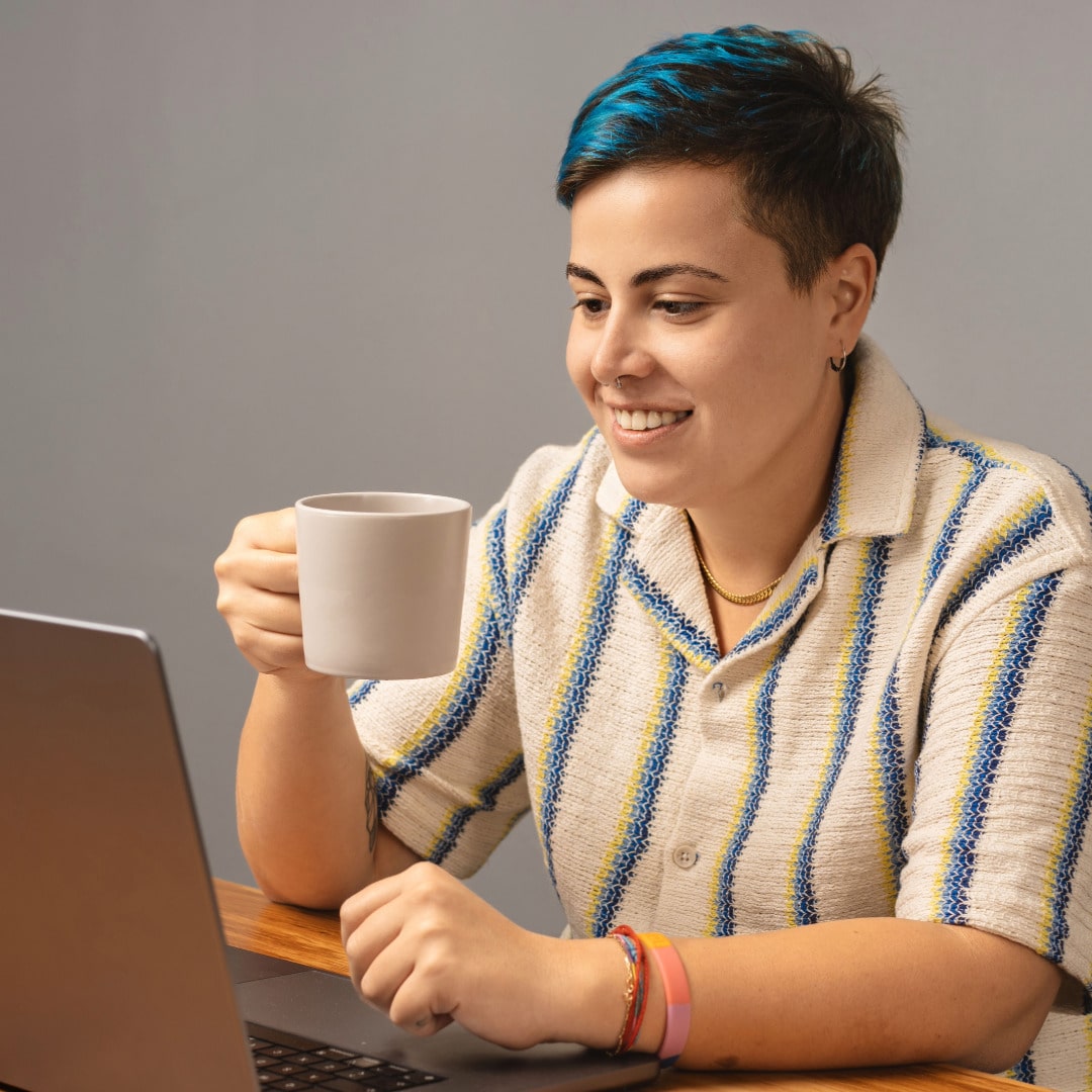 Nonbinary person with short dark hair and blue highlights wearing a blue and yellow striped cream button down and two bracelets drinking a hot beverage from a cream-colored mug smiling while working with their out-of-network therapist.