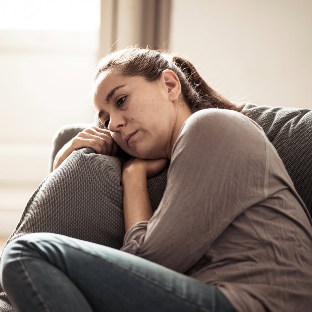 Young woman with her hair pulled back in a pony tail wearing a brown long sleeve shirt and jeans curled up on the couch trying to overcoming illness-related anxiety.