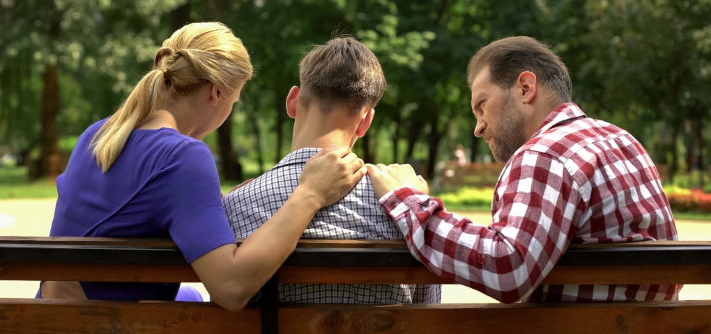 Parents sitting on either side of their teen son with their hands on his shoulders on a park bench facing trees, looking concerned and supportive after working with a parenting counseling therapist.