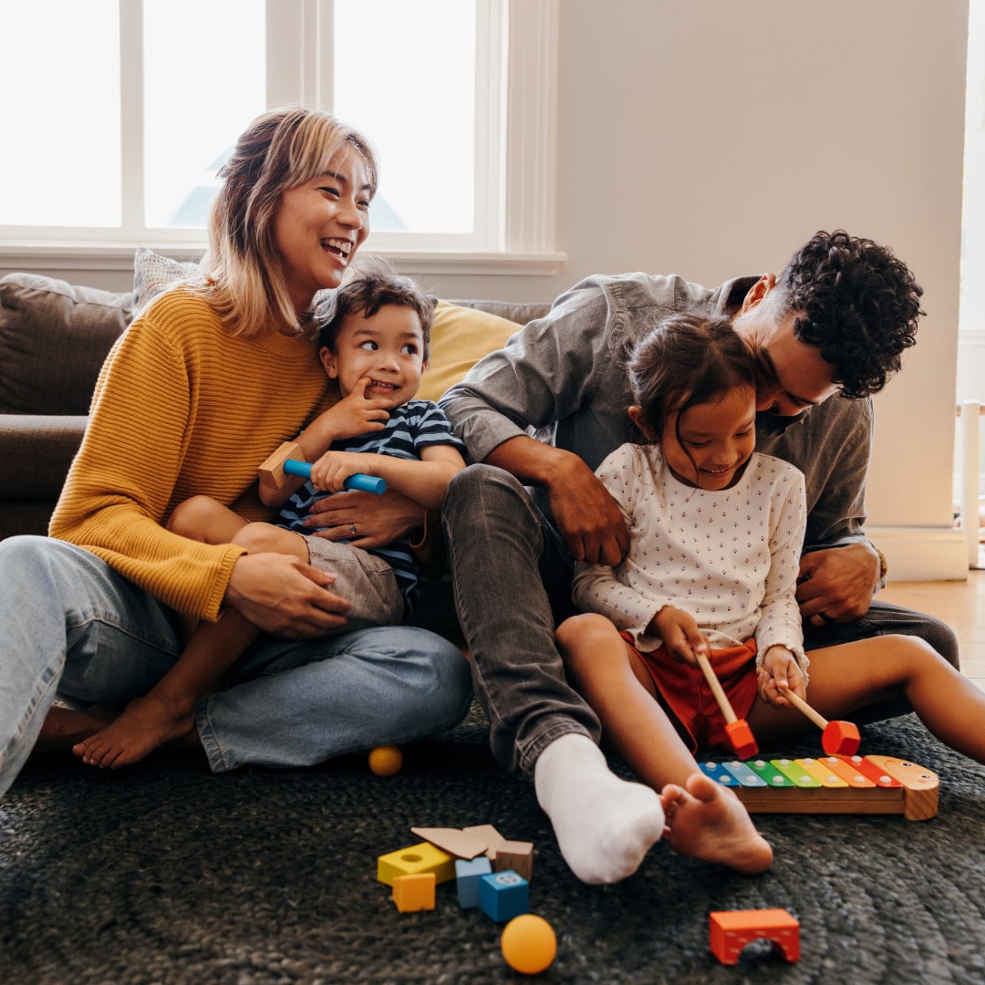 A multiracial family sitting on the floor together in front of the couch. Mom is holding the younger son while Dad sits behind the daughter, whispering in her ear. This family has benefited from parenting counseling.