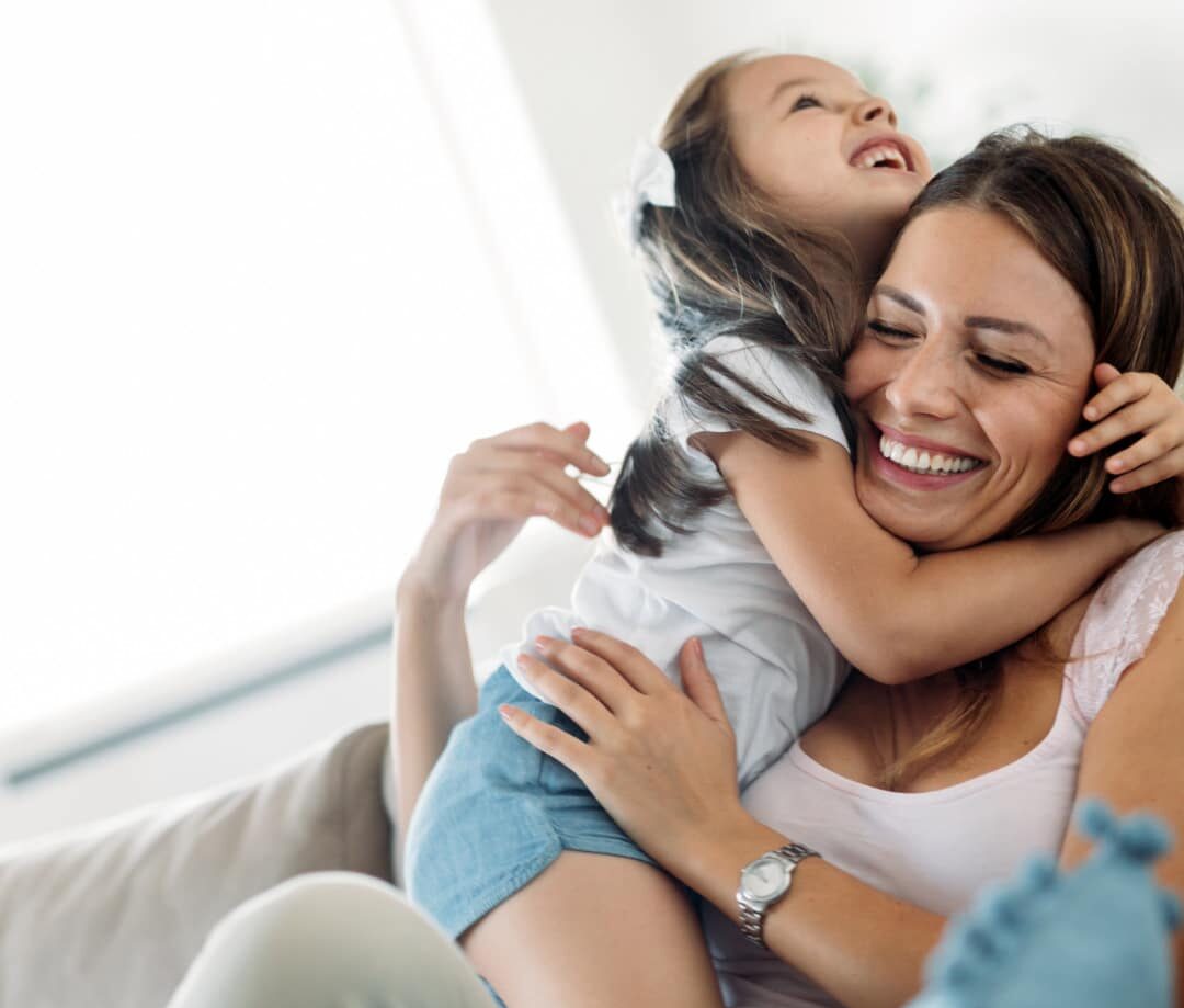 A woman with long dark hair wearing a white tank top and tan pants sitting on a couch smiling holding her 5 year old daughter who also has long dark hair, wearing a white short sleeve t-shits and light blue shorts and is looking up wtih her chin on mom's head smiling. They are both having fun after mom has found some strategies to cope with her PDA.