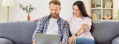 A couple sitting close together focused on the laptop on the man's lap while getting personalized parenting support from an online parenting therapist to improve parenting skills.