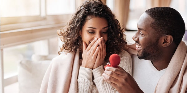 A black couple sitting on a couch with the man presenting a ring in a red velvet box while the woman holds her hands over her mouth in excitement ready to start premarital therapy online.