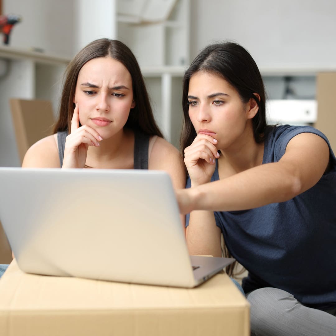 Partners deciding to see an out-of-network therapist to protect their private health records due to chronic illness. Both have long, straight dark hare and are sitting in front of a laptop on a table. The person on the left is wearing a gray tank top and leaning her chin on her fingers. The person on the right is wearing a dark blue short sleeve t-shirt and resting her chin on her curled hand while pointing to the computer screen with her other hand.