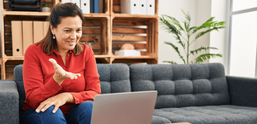 A middle aged woman with short dark hair wearing a red long sleeved shirt and jeans seeing a therapist online from her office smiling and gesturing about feeling understood and accepted.