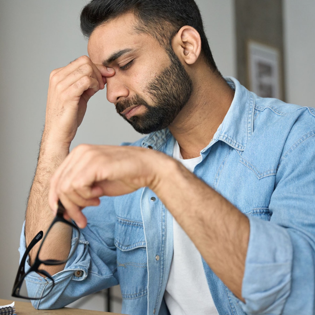A man in a light blue shirt with his glasses in his hand rubbing his face and showing signs of caregiver stress.