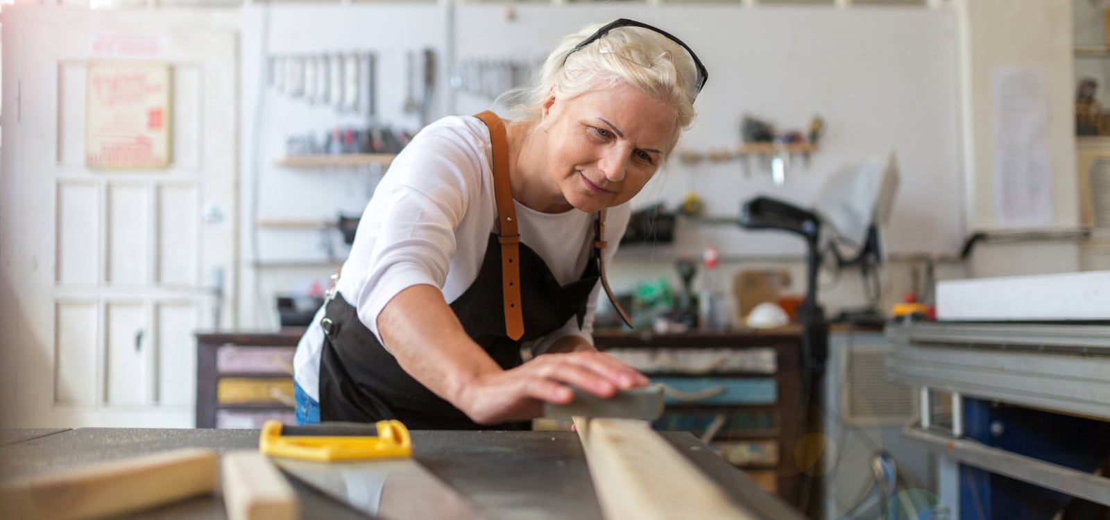 Woman in her 60s with her hair pulled back, wearing a shop apron bending over a shop table working with a piece of wood using strategies for dealing with PDA by focusing her woodworking interest .