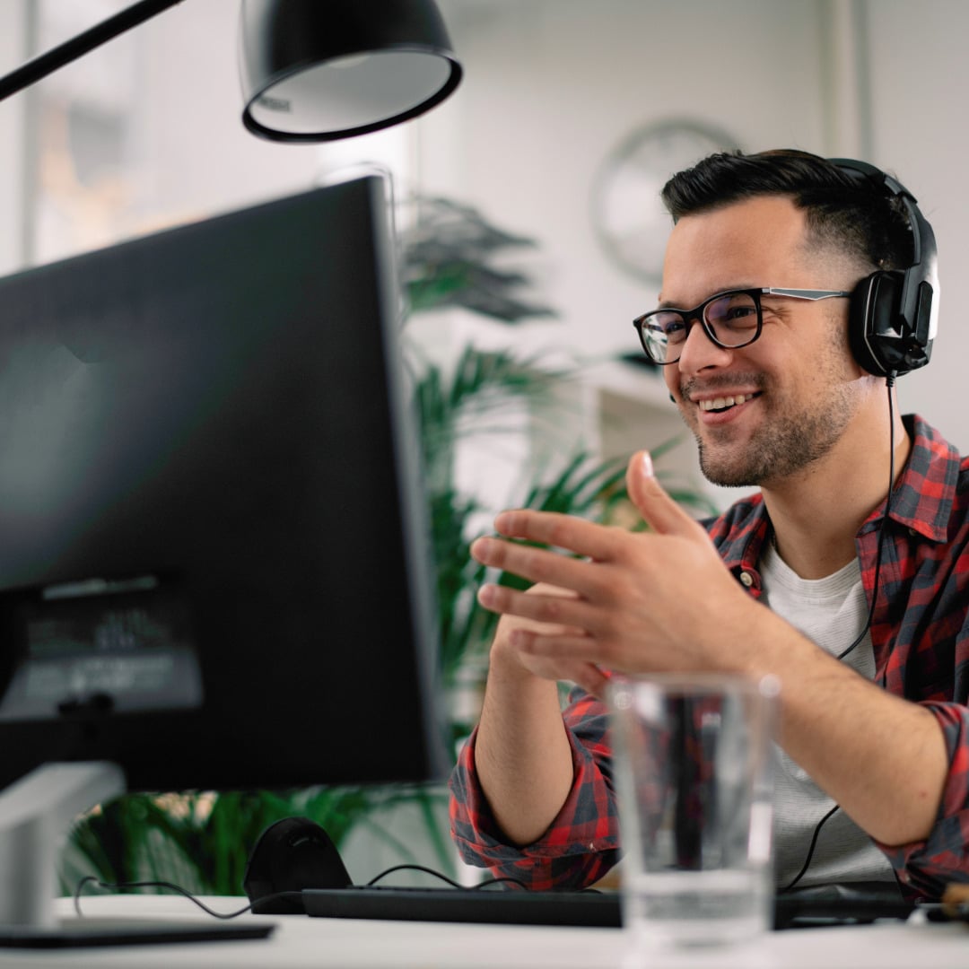 Neurodivergent guy with short, dark hair and glasses wearing a red plaid shirt over a white t-shirt and headphones attending a therapy session online with a clear, almost empty glass of water in the foreground..