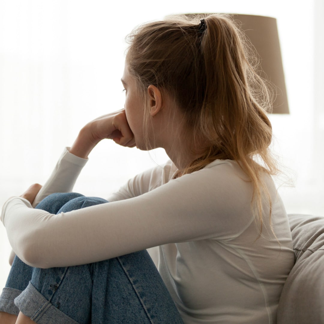 Teen girl with blond hair in a ponytail sitting on a beige couch with her arm around her knees and her chin resting on her hand looking away out the window thinking about understanding addiction.