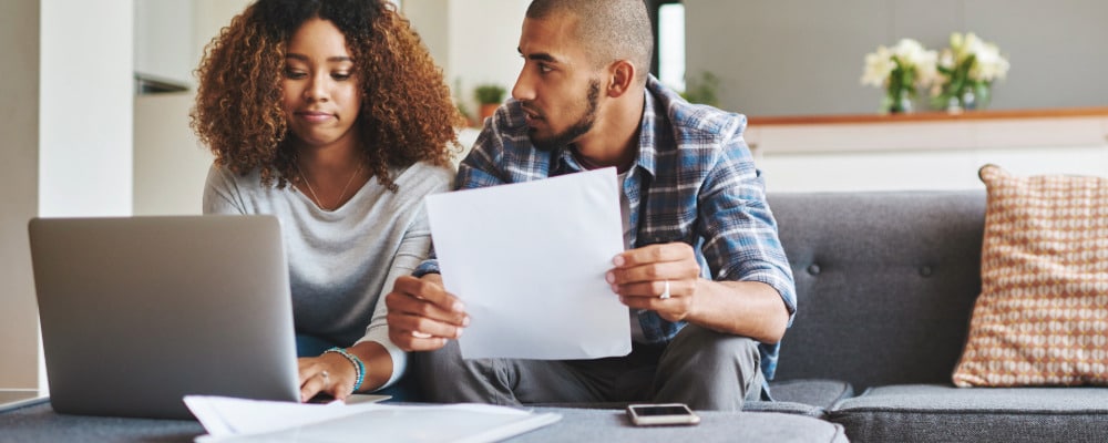 A confused couple working on understanding different types of health insurance sitting on the couch together going over mental health insurance paperwork with a laptop.