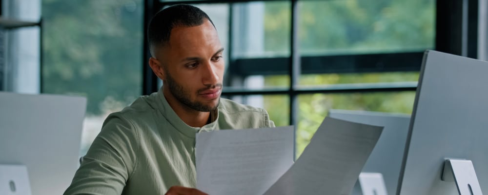 Young Black man with short dark hair sitting in front of his laptop with paperwork working on using out-of-network benefits for mental health services.