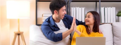 A couple celebrating a parenting success. The wife is sitting on a white couch with the laptop. The husband is behind the couch, leaning on the back with his arm around his partner while they high-five during their virtual counseling for parents session. 