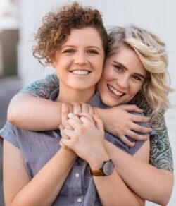 Queer couple embracing outside in front of a white wall after wondering what to expect in premarital counseling.